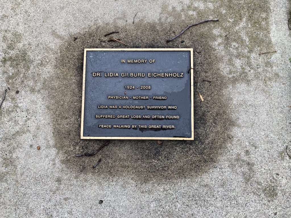 a plaque embedded in the sidewalk with the inscription: In memory of Dr. Lidia Gilburd Eichenholz 1924-2008 Physician Mother Friend Lidia was a holocaust survivor who suffered great loss and often found pleace walking by this great river.