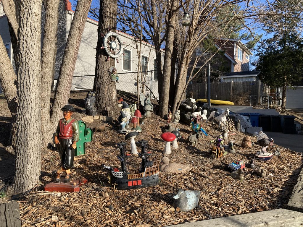 dozens of figurines displayed at the edge of a yard, including an owl, a gnome, several mushrooms, and the helm of a ship hanging from a tree.