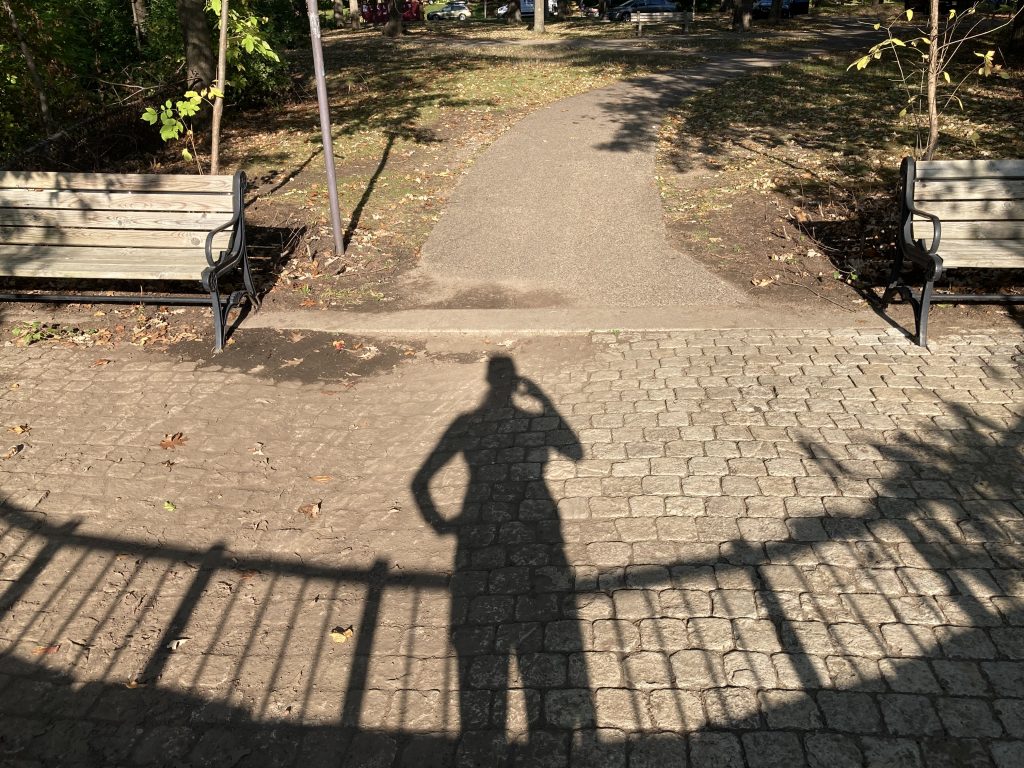 The shadow of a runner and the railing from an overlook above the Mississippi River. One hand of the shadow is holding a phone to her face, the other is perched on her hip. 