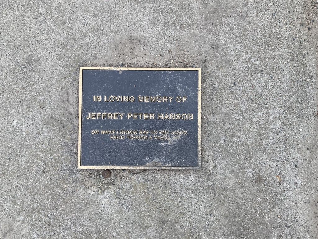 A plaque near a bench. It reads: In Loving Memory of Jeffrey Peter Hanson. "Oh what I would say to you again" from Losing a Year